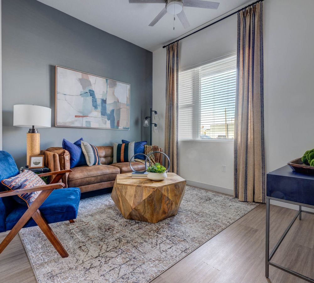 Interior of a modern living room at Alta Stone at The Gateway, featuring a tan leather sofa with blue accent pillows, a blue velvet chair, a geometric wooden coffee table, and a floor lamp with a wooden base.